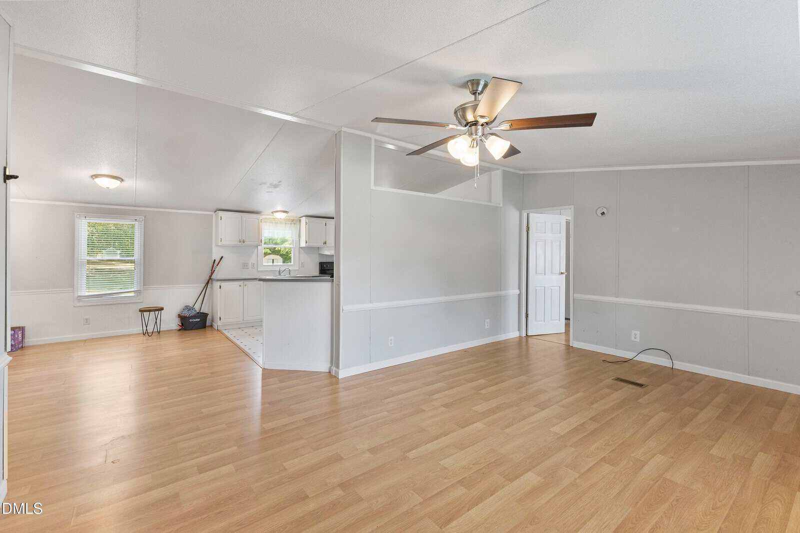 Open living area with light wood floors, gray walls, ceiling fan, and white trim. Kitchen with white cabinets and appliances is visible in the background. Large windows provide natural light.