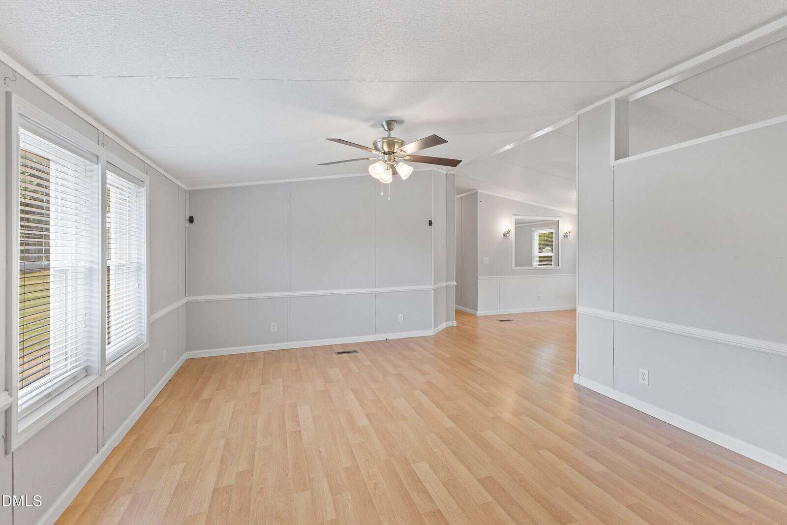 Spacious empty room with light wood flooring, gray walls, white trim, a ceiling fan with lights, several windows, and an open entryway to another room with similar decor.