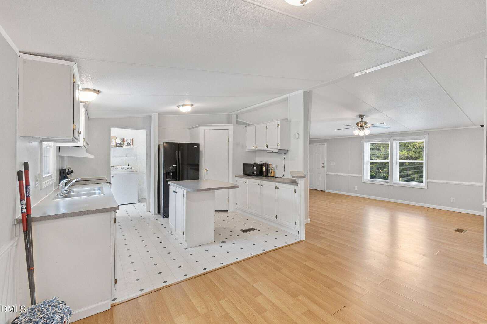 Open kitchen with white cabinets, black appliances, small island, and an adjacent laundry area. To the right, a spacious living room with wood flooring, ceiling fan, and large windows letting in natural light.