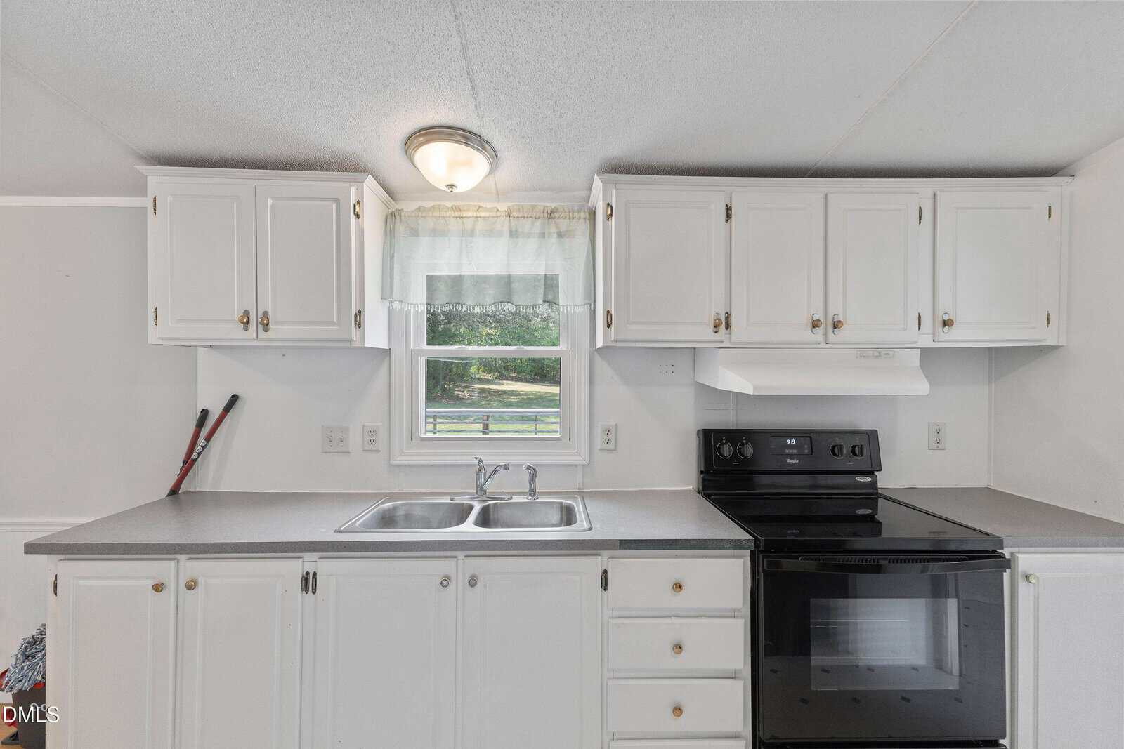 A kitchen with white cabinets, a double sink under a window, a black electric stove with oven, and a gray countertop. Two brooms are leaning in the corner. Natural light comes through the window.