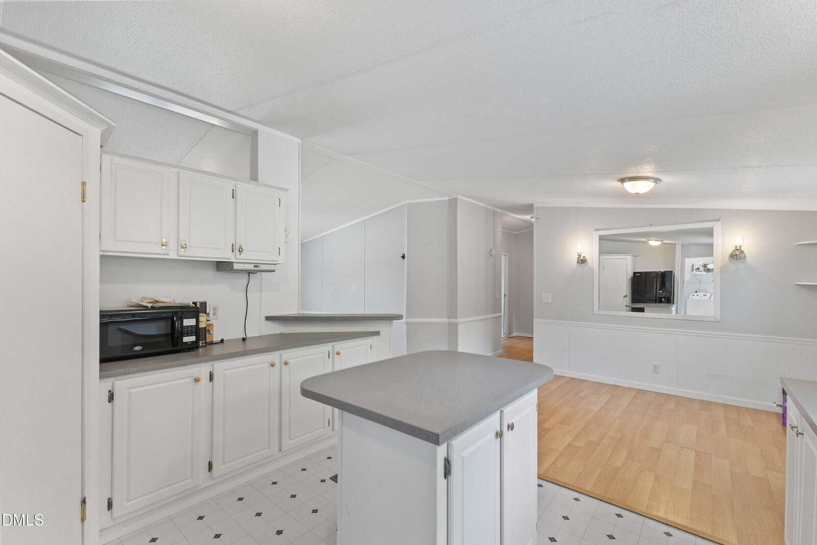 Bright kitchen with white cabinets, a gray countertop island, black microwave, and open layout leading to a living area with light wood flooring, wall mirror, and built-in shelves.