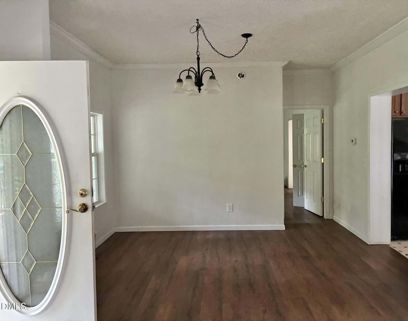 A mostly empty room with light walls, wood floors, a three-light ceiling fixture, an open white door with glass, a window, and two open doorways leading to other rooms.