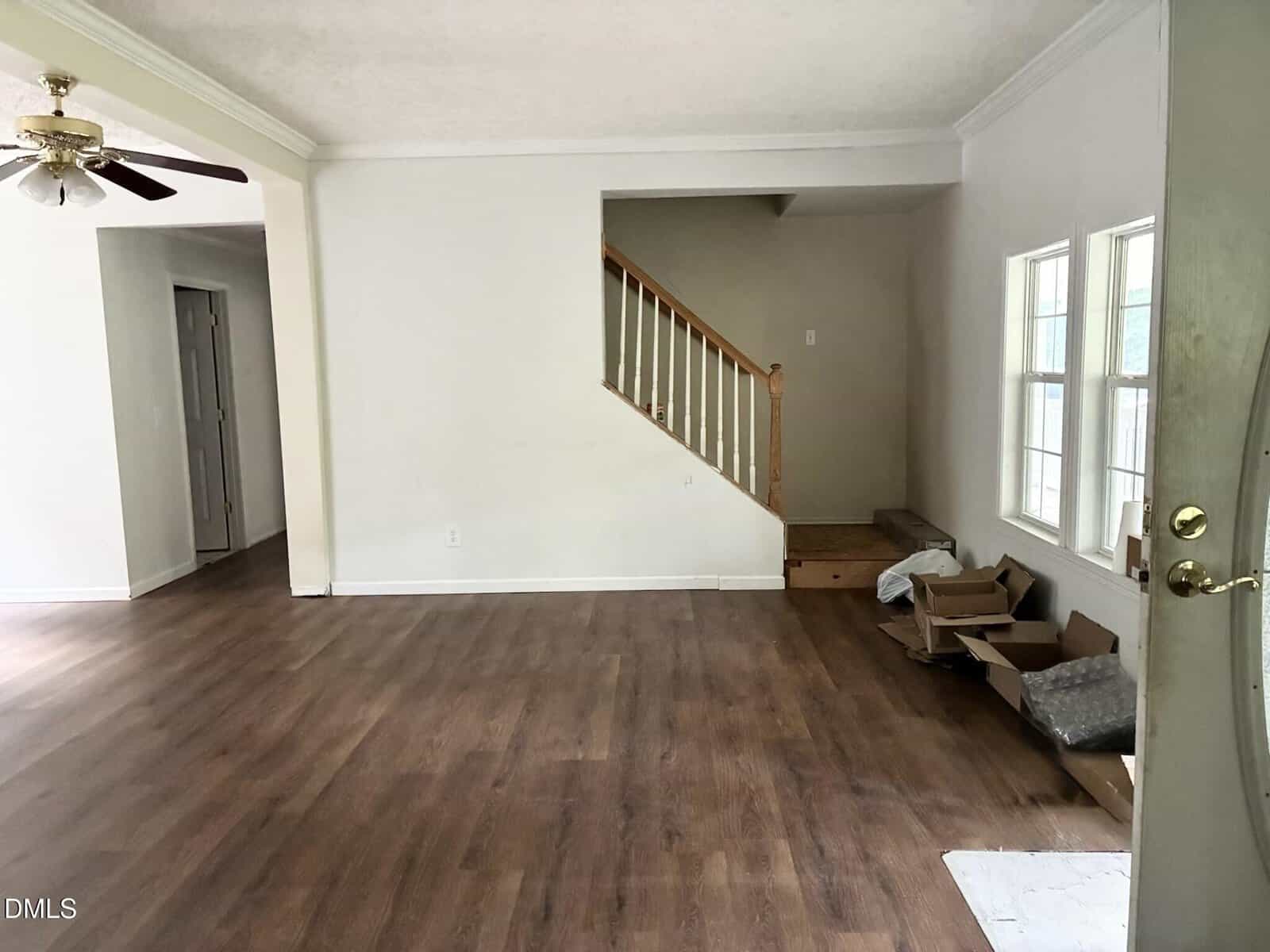 Unfurnished living room with wooden floors, white walls, and a staircase with a wooden railing. Three windows let in light. Several unpacked boxes are on the floor near the windows and door.