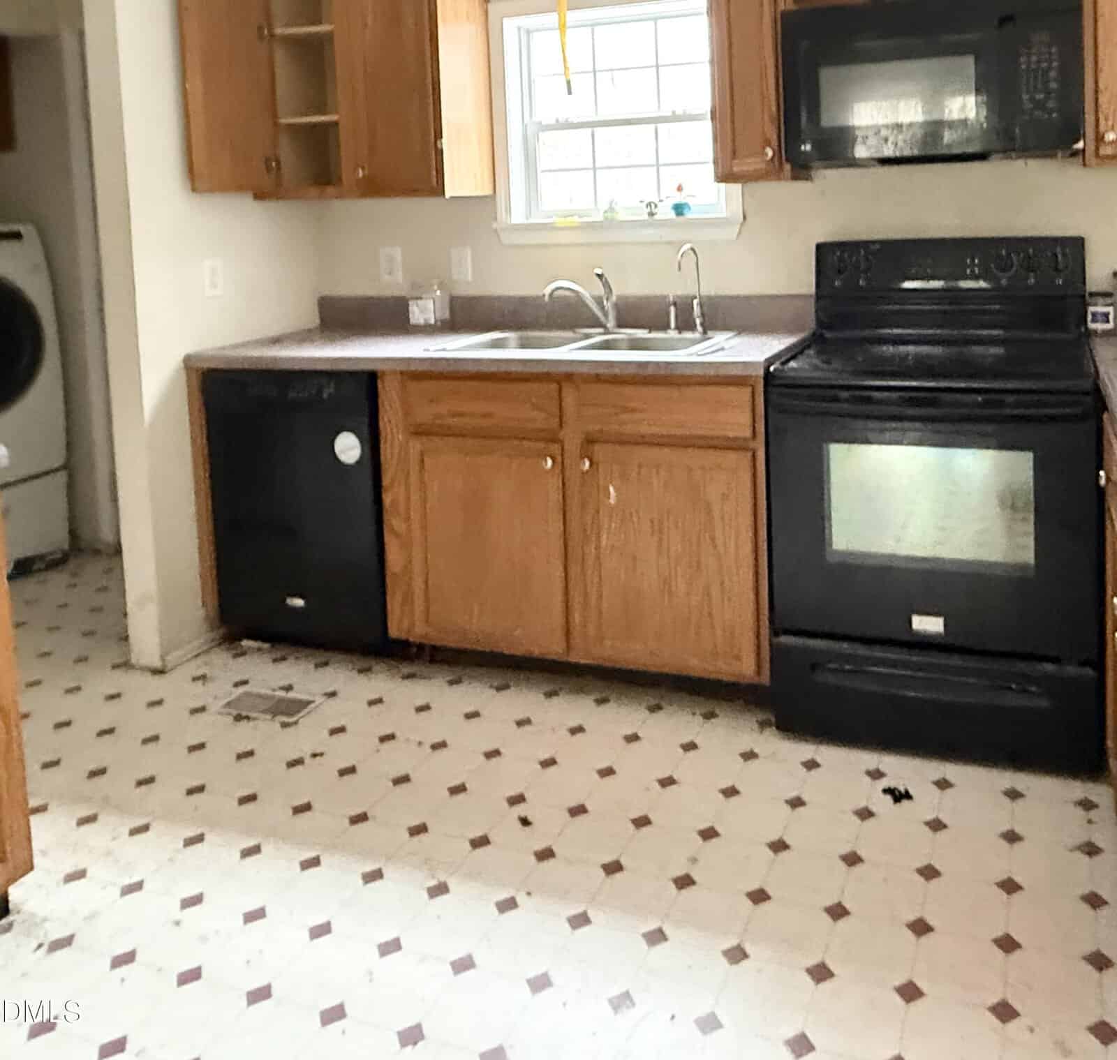 A kitchen with wooden cabinets, black appliances including a stove and dishwasher, a double sink under a window, and a visible laundry area with a washing machine in the adjacent room. The floor has a white and red tile pattern.