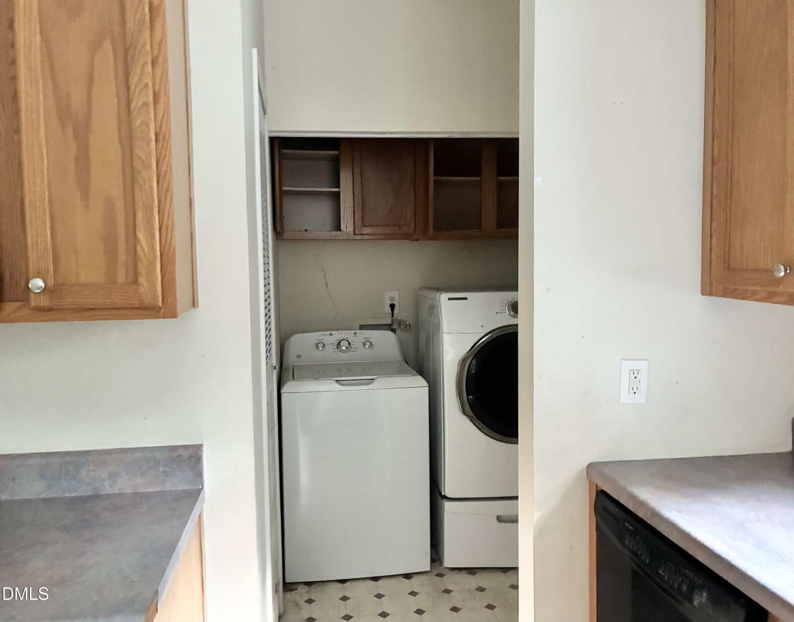 A small laundry nook with a washing machine and dryer, wooden cabinets above, and light-colored walls. Kitchen counters and cabinets are visible in the foreground.