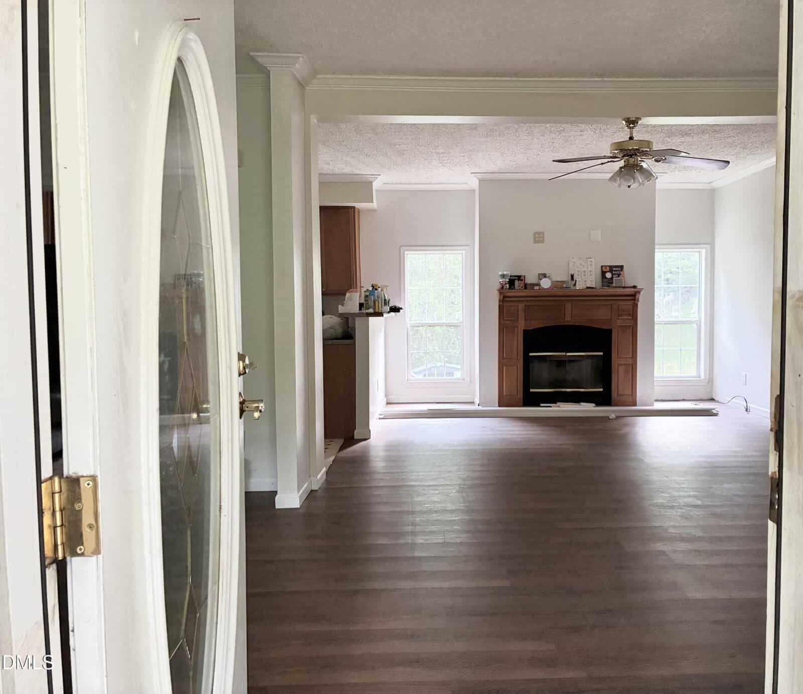 View from an open front door into a living room with wood floors, a ceiling fan, and a brick fireplace. Large windows let in natural light; the kitchen area is partly visible to the left.