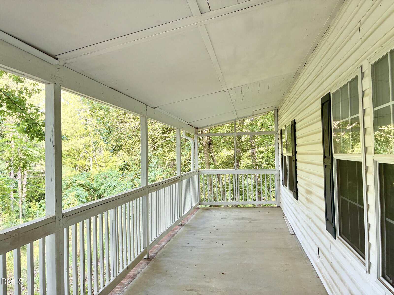 Covered porch with white railing and siding, black shutters, and large windows. The porch overlooks green trees and has a sloped roof and concrete floor. Bright natural light fills the space.
