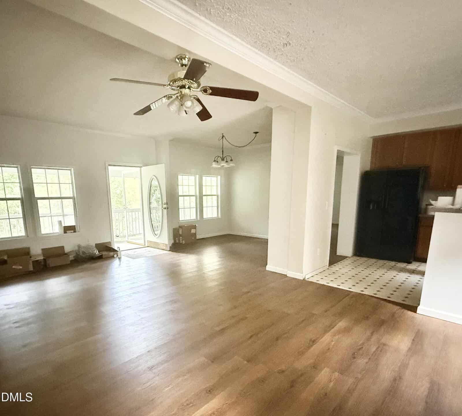 Spacious, unfurnished living area with wood floors, white walls, ceiling fan, large windows, and an open kitchen with tiled floor and black appliances. Boxes are stacked near the windows and door.
