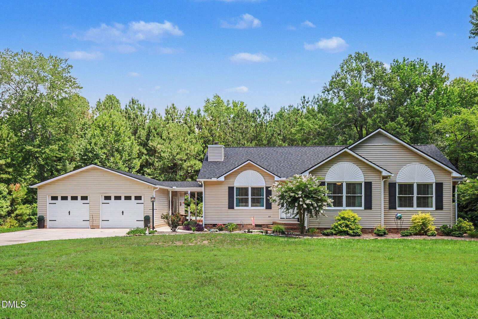 Charming single-story beige house at 4070 Raney Way Drive in Stem, featuring an attached three-car garage, green shutters, and a classic brick foundation. The landscaped front yard and large lawn are surrounded by trees, all beneath a clear blue sky with scattered clouds.