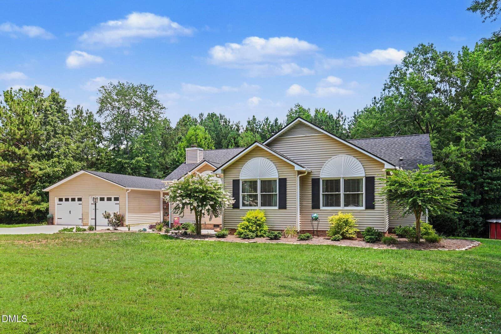 Single-story beige house with white trim, dark roof, large windows, and attached garage. The yard is grassy with trees and shrubs, set against a backdrop of dense greenery under a blue sky with scattered clouds.
