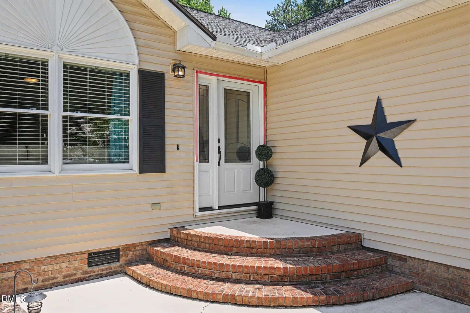 Front entrance of a house with beige siding, a white door, brick steps, a large black star decoration on the wall, and two potted topiary plants beside the door.