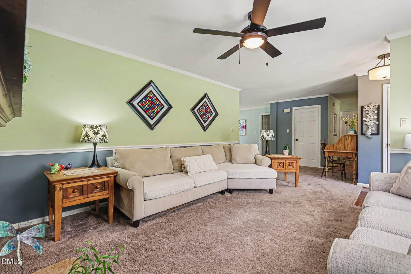 Living room with beige sofas, wooden side tables, lamps, ceiling fan, and green accent wall displaying two framed artworks. Dining area and hallway visible in the background. Carpeted floor throughout the space.