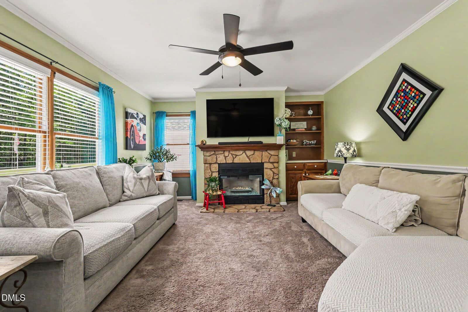 A cozy living room with two sofas, a stone fireplace, TV above it, bookshelves, green walls, blue curtains, a ceiling fan, and carpeted floor. Plants and framed art decorate the space.