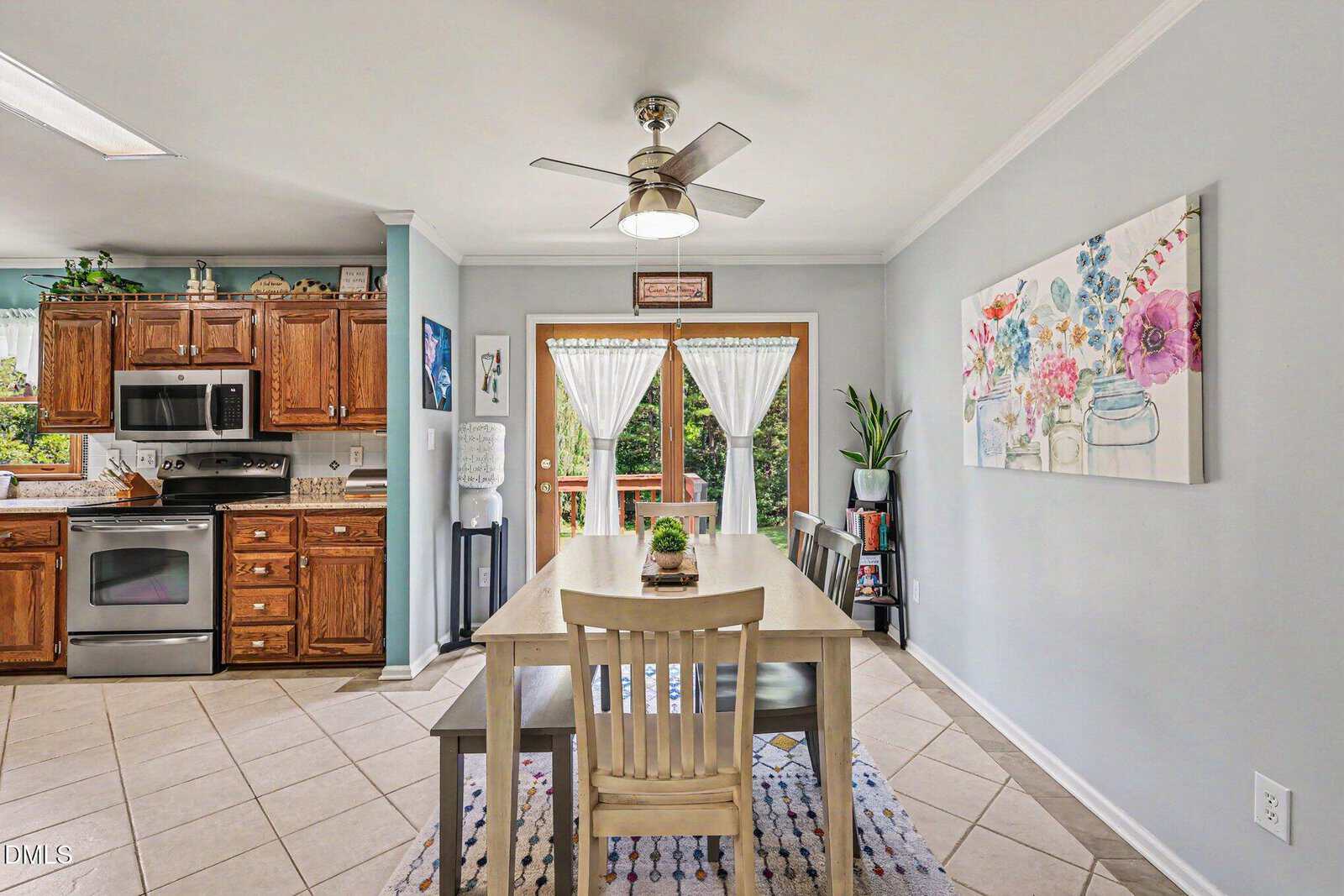 Bright kitchen with wooden cabinets, stainless steel appliances, a central dining table, ceiling fan, wall art, indoor plants, and glass doors leading outside with sheer white curtains.