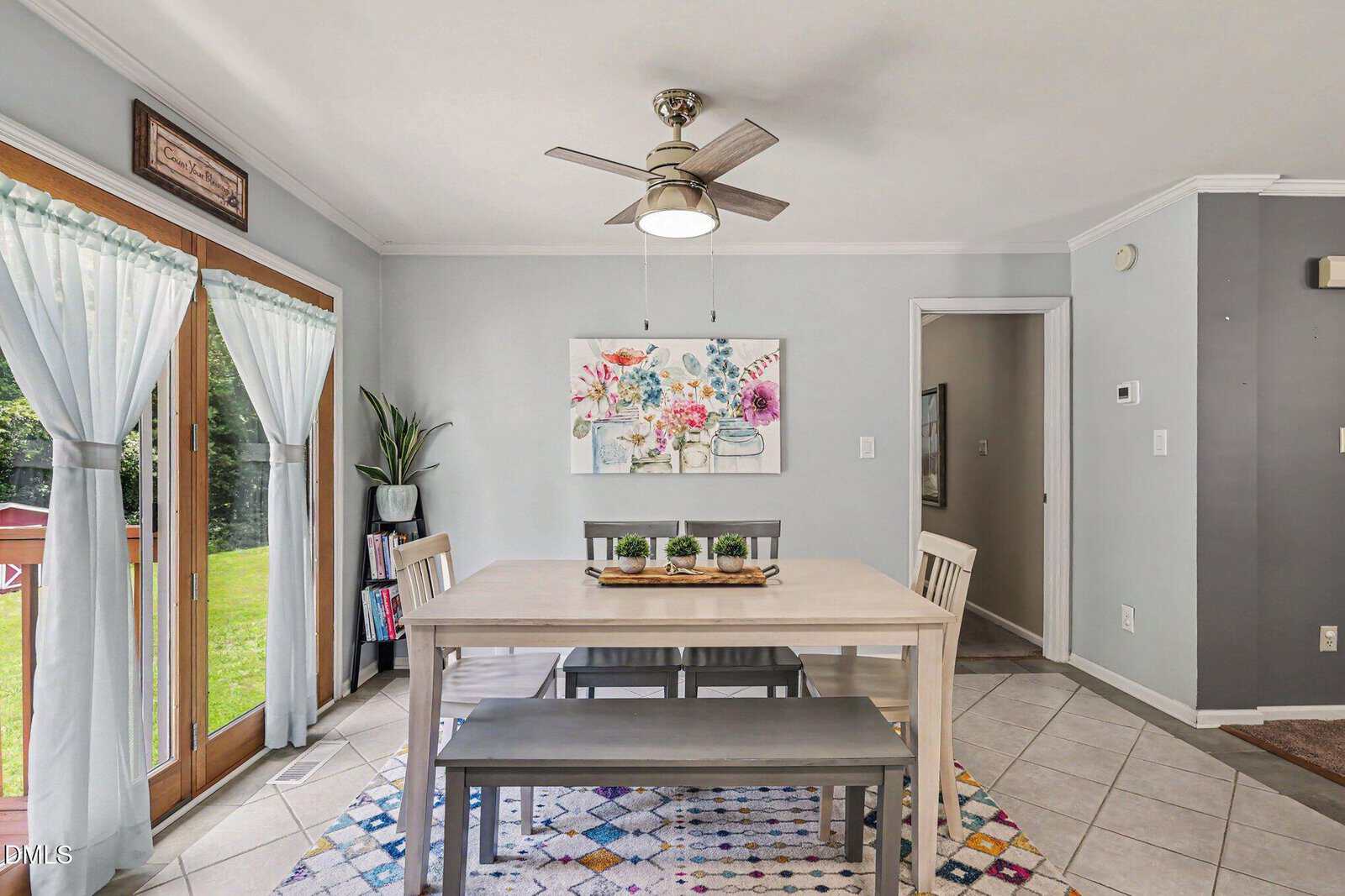Bright dining area with a wooden table, bench, and chairs on a patterned rug. Large glass doors with sheer curtains let in natural light. A ceiling fan hangs above, and there’s floral art on the wall. Plants decorate the room.