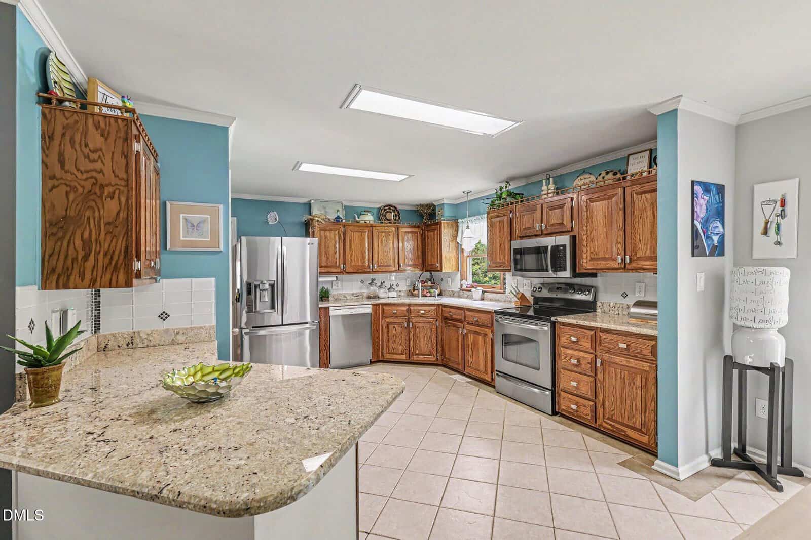 Spacious kitchen with wood cabinets, stainless steel appliances, granite countertops, light tile floor, teal accent wall, and decorative items on shelves and counters. Large window over sink lets in natural light.