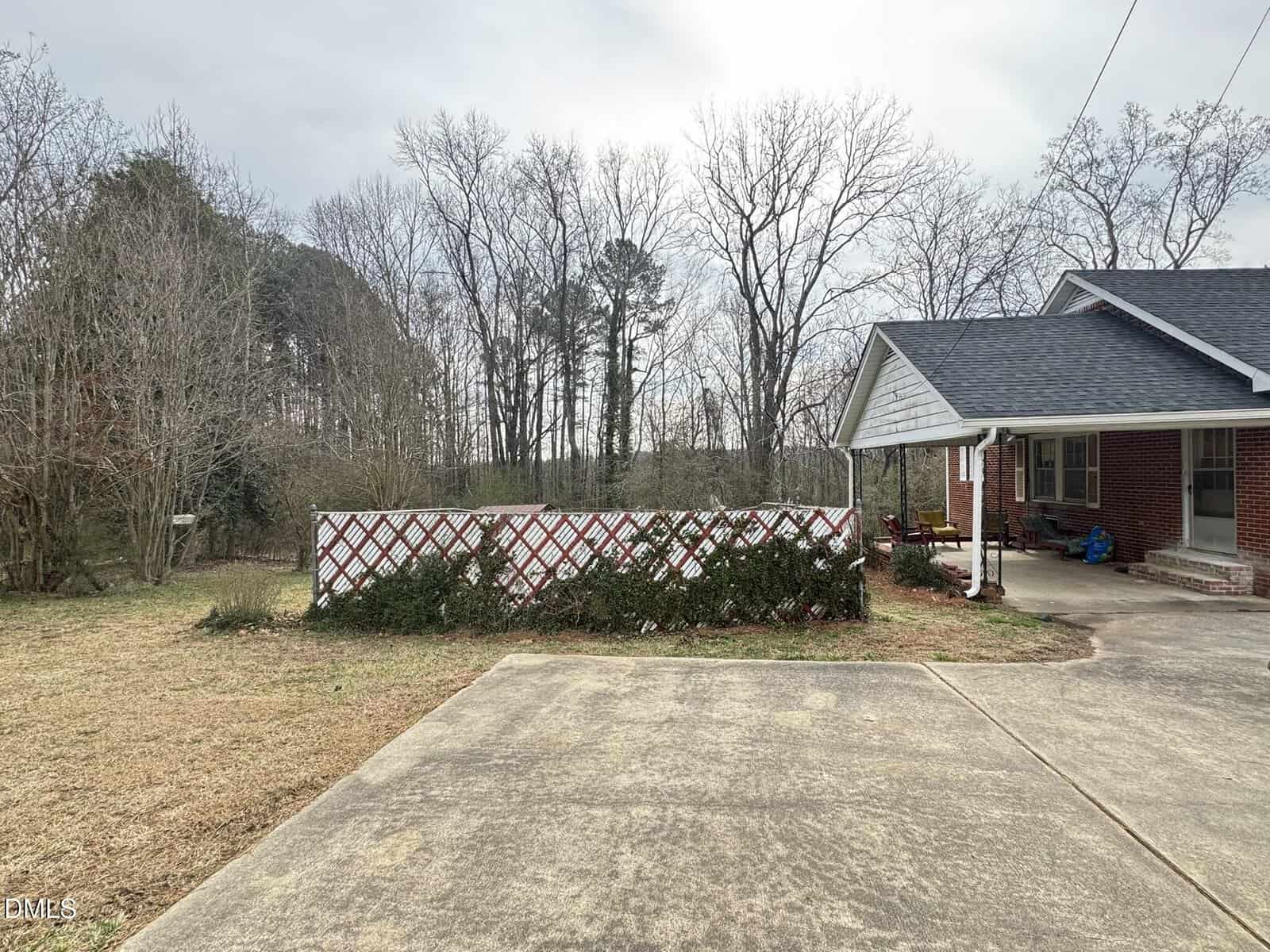 Brick house with front porch, driveway, and a white lattice fence covered partly by shrubs. Set along 580 US Hwy 158 Bus E in Warrenton, this home is surrounded by leafless trees and bushes filling the background under a cloudy sky.