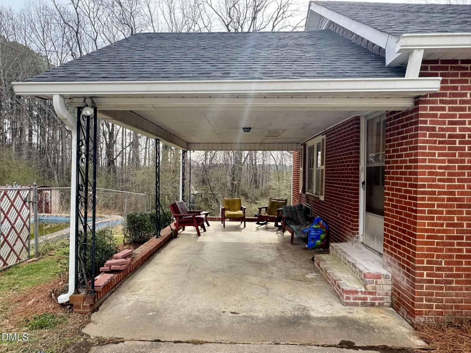 A covered patio attached to a red brick house with several outdoor chairs, a small table, and some gardening supplies. Leafless trees and a fenced yard are visible in the background.