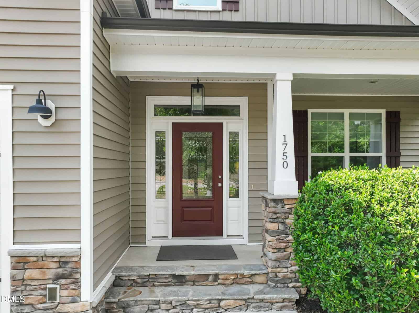 A house entrance with a red front door, white trim, stone steps, a large window, and a porch light. House number 1750 is on a white column next to green bushes.