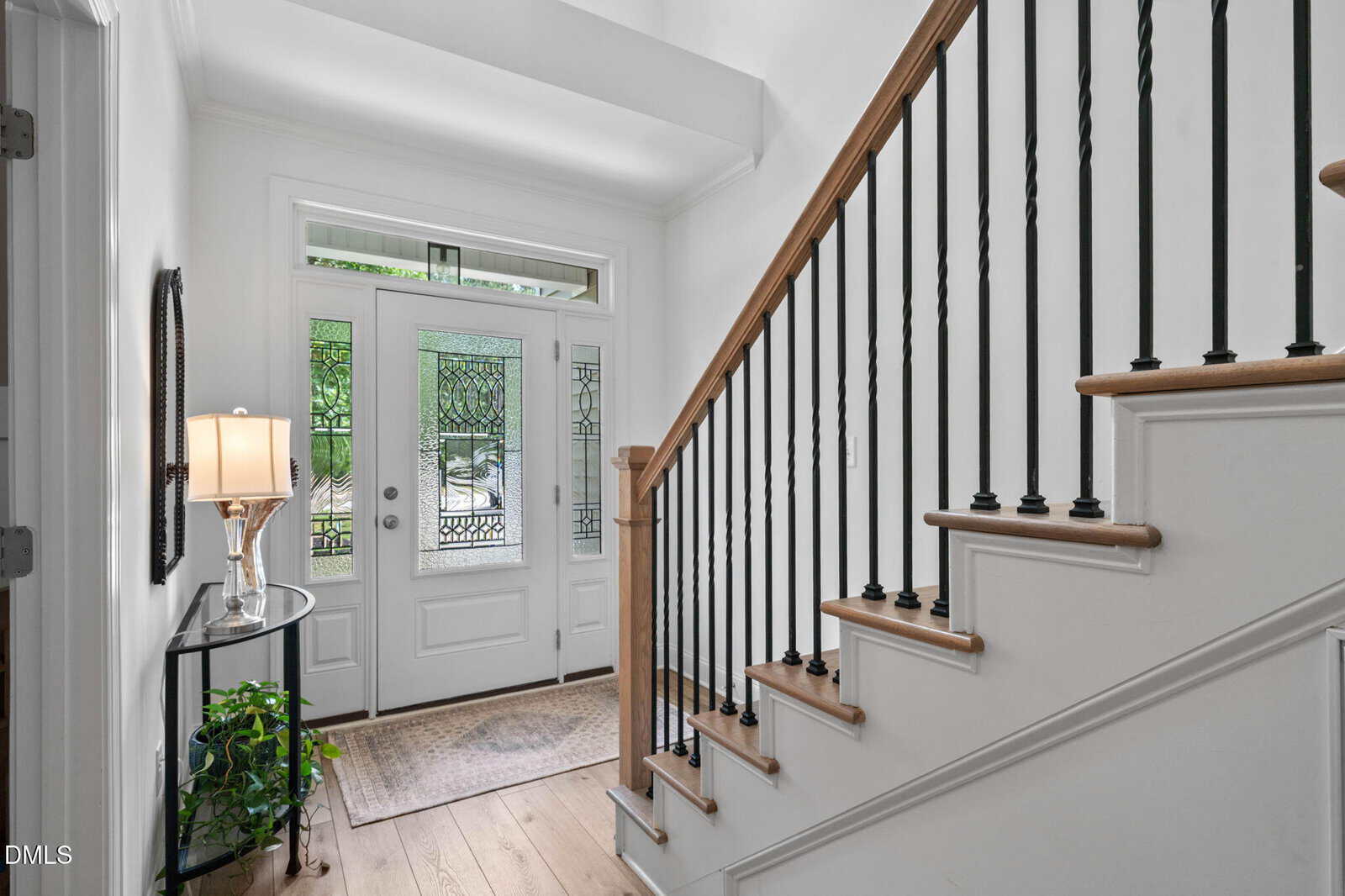 Bright entryway with a glass front door, intricate glass panels, a side table with a lamp and plant, and a wooden staircase with black metal balusters. Light wood floors and white walls create an airy, welcoming feel.