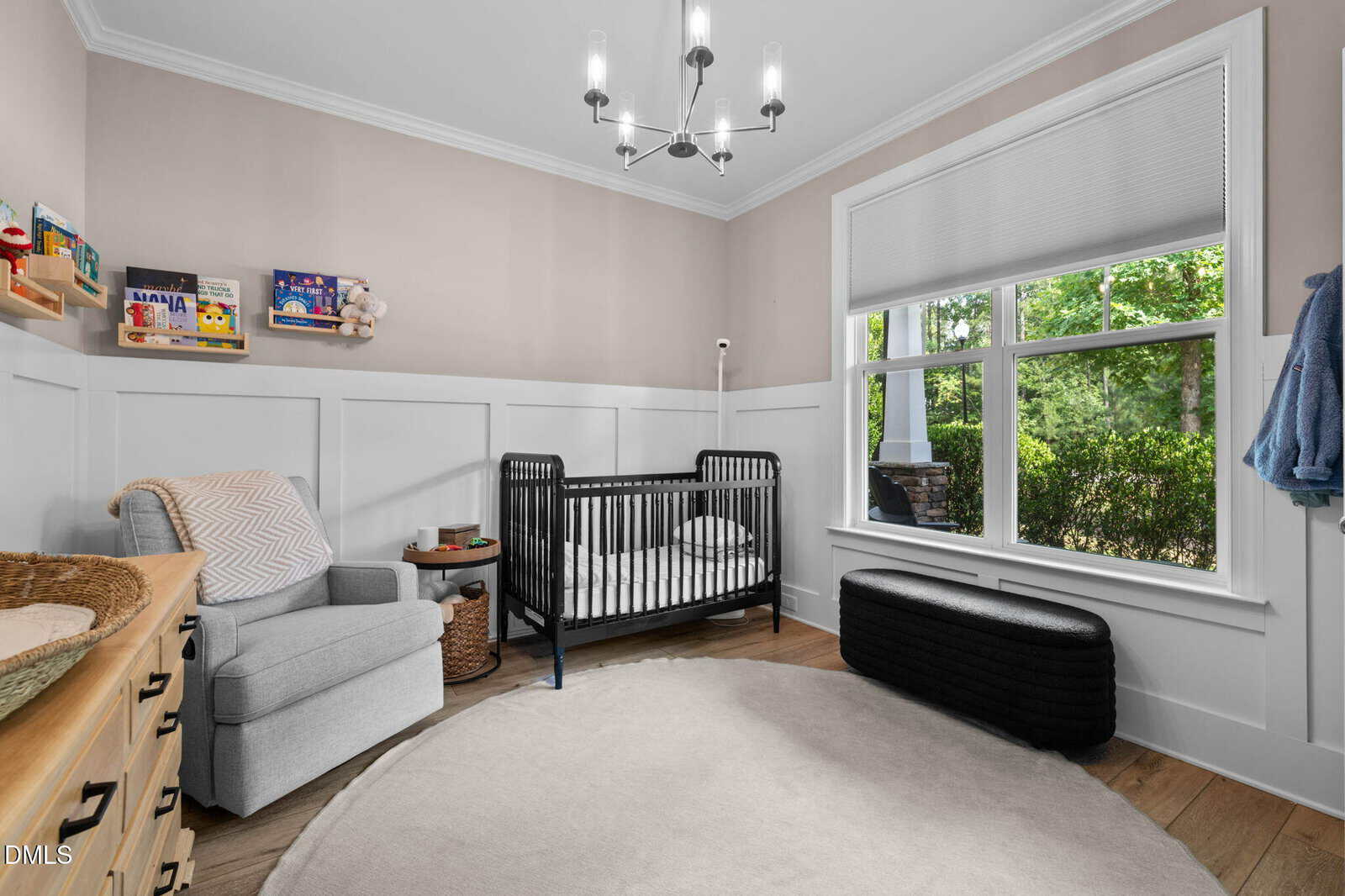 Neutral nursery with a black crib, gray armchair, round rug, dresser with baskets, books on wall shelves, window seat bench, and large window showing greenery outside. Modern light fixture on ceiling.
