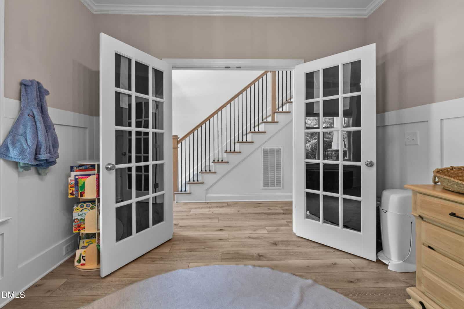 A room with open French doors leading to a staircase. Toys and books are on a shelf to the left, a blue robe hangs on the wall, and a dresser and trash can are to the right. Light wood flooring throughout.