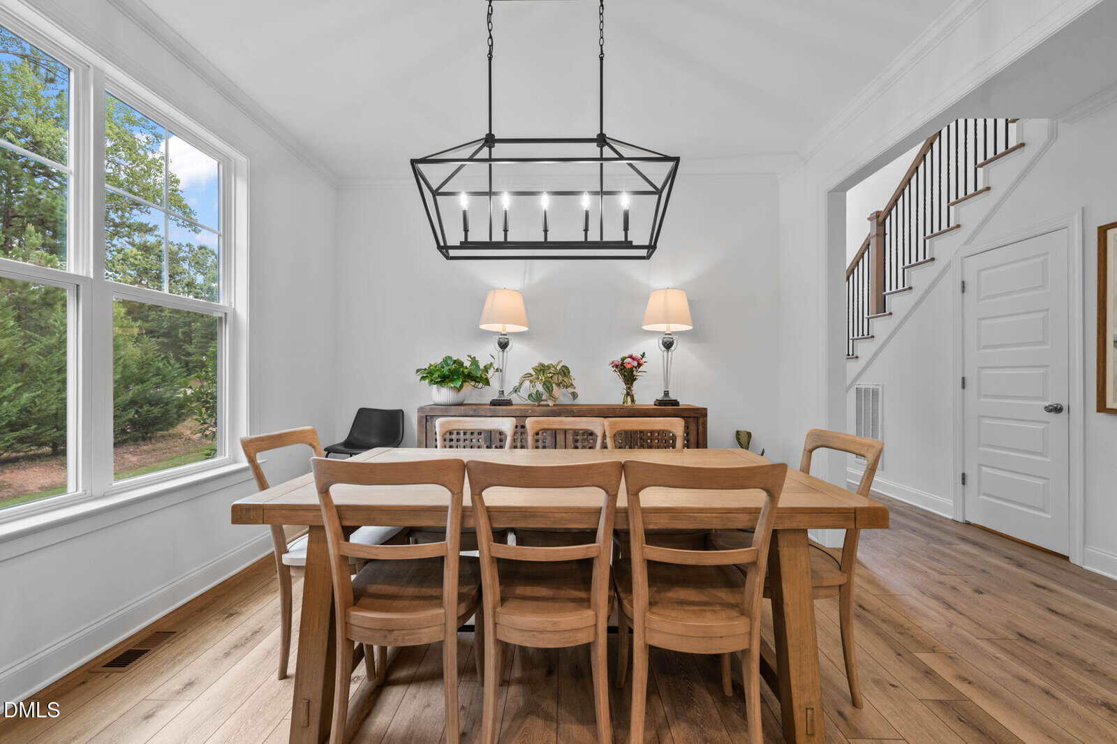 Bright dining room with a wooden table and eight chairs, modern light fixture, sideboard with lamps and plants, large window, and wooden floors. Stairs and a door are visible in the background.