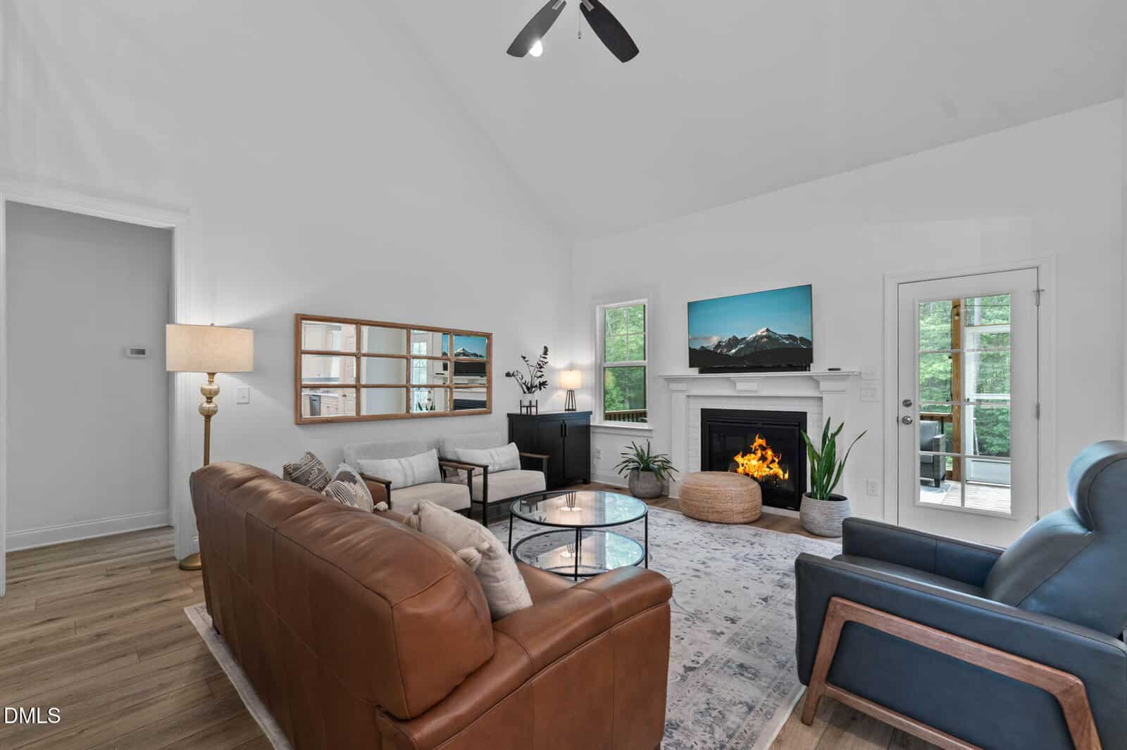 Modern living room with brown leather sofa, blue armchair, fireplace with a fire, TV displaying mountains, large windows, glass door to outside, and neutral decor on a patterned rug. Cozy and well-lit space.