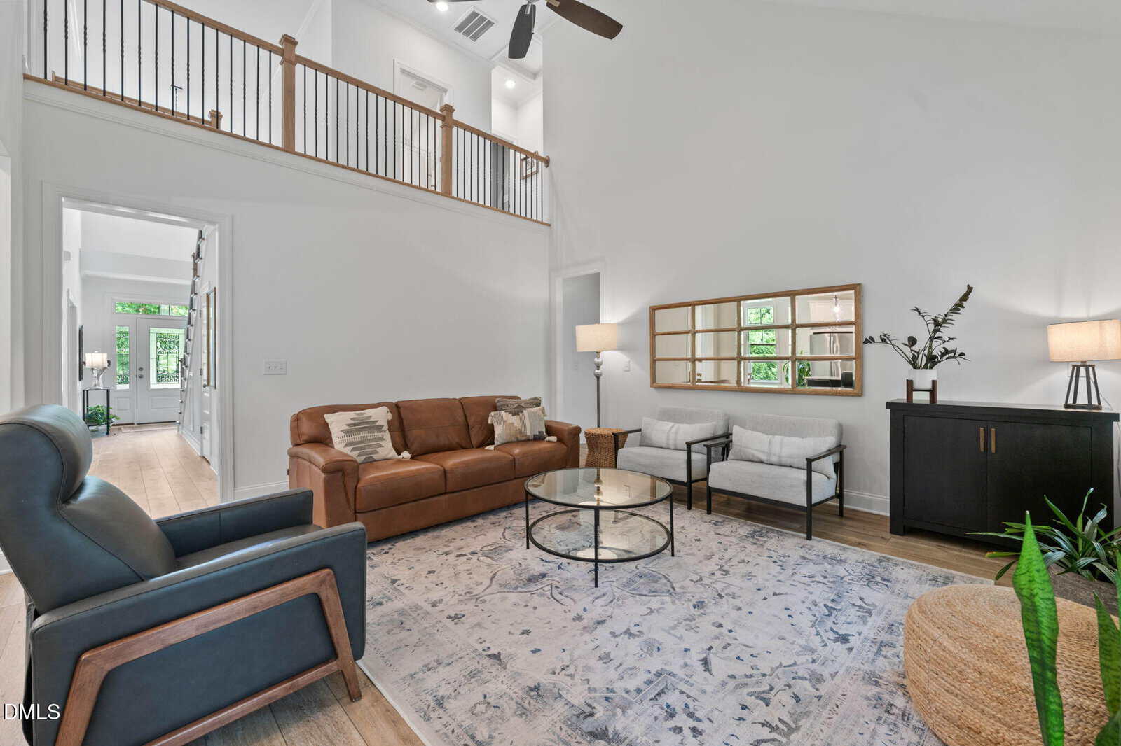 Modern living room with a brown leather sofa, two gray armchairs, round glass coffee table, large rug, black sideboard, mirror, plants, and wood flooring. Loft railing and high ceilings visible above.