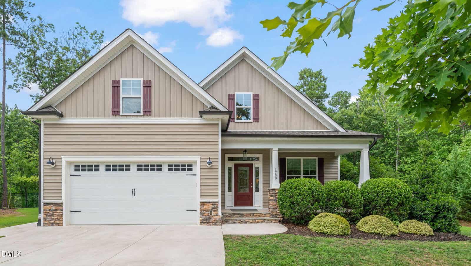 A beige two-story house with white trim, a covered front porch, maroon shutters, and a two-car garage is nestled at 1750 River Club Way in Franklinton. The beautifully landscaped yard features green bushes and trees surrounding the home, creating a welcoming and peaceful atmosphere.