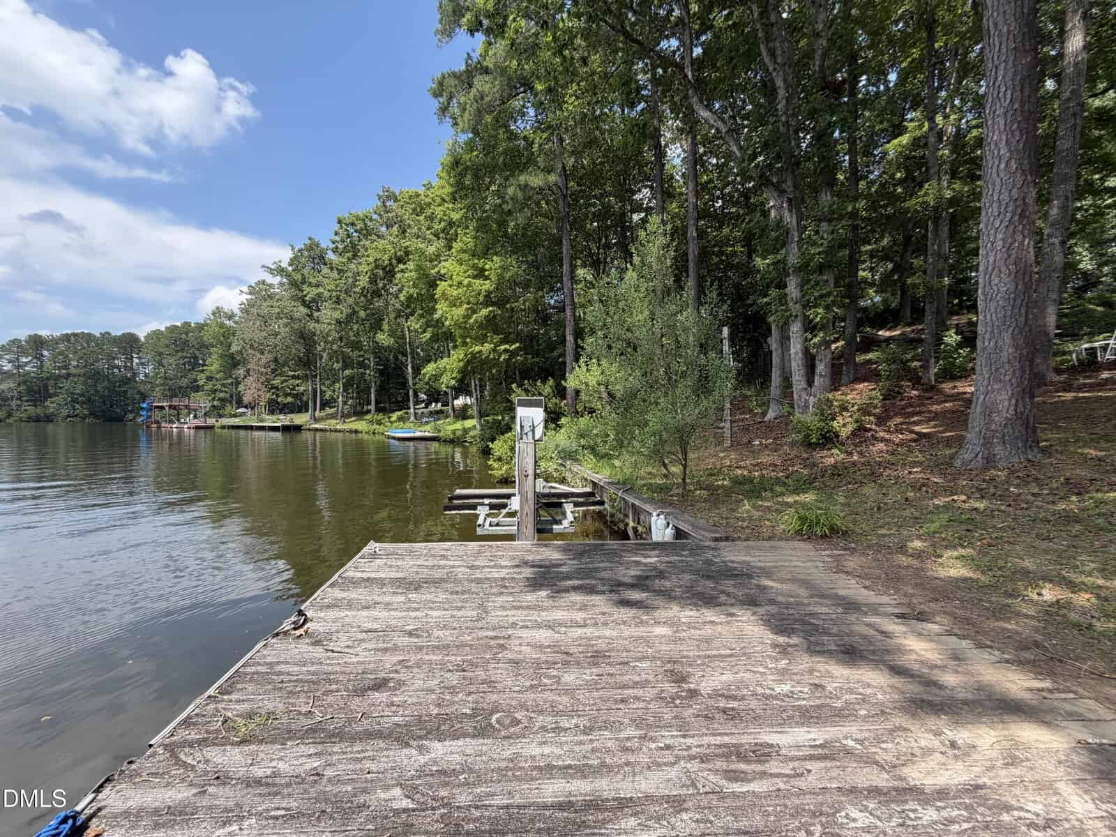 A wooden dock extends over a calm lake, surrounded by trees and greenery. The shoreline curves left, and boats are visible in the distance. The sky is partly cloudy.