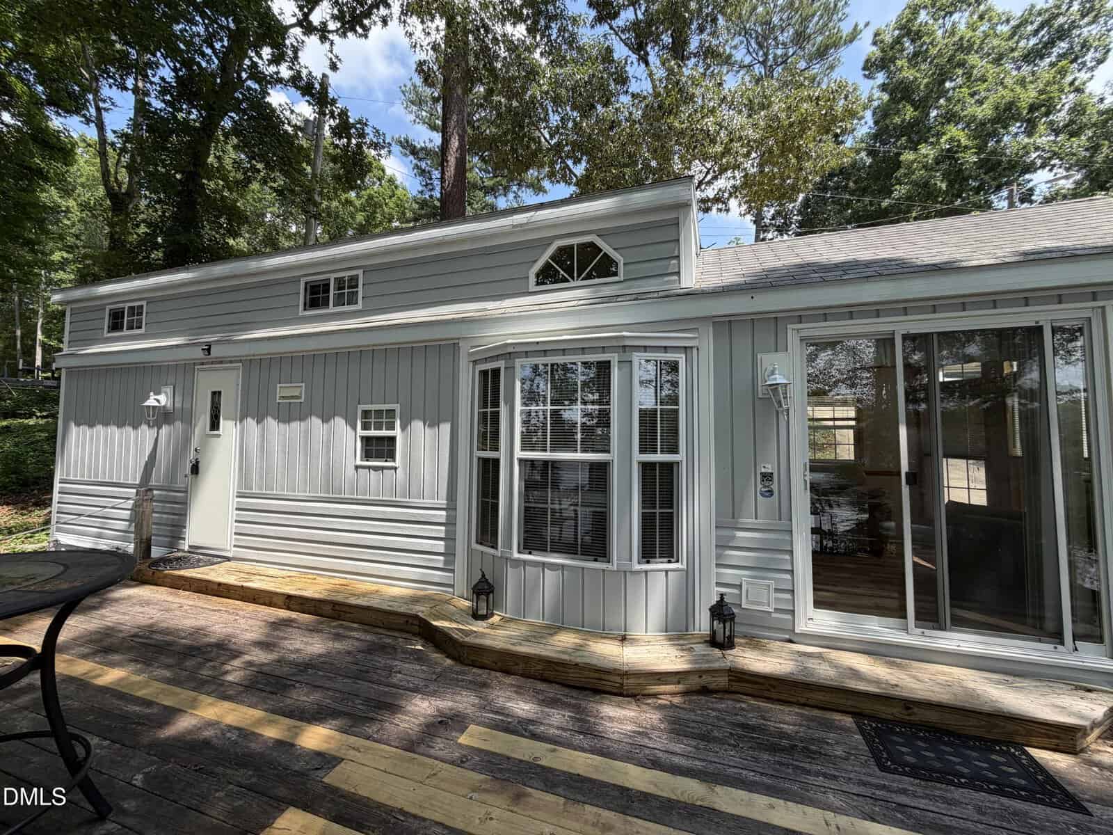 A small gray house with large windows, white trim, and a wooden deck surrounded by tall trees on a sunny day.