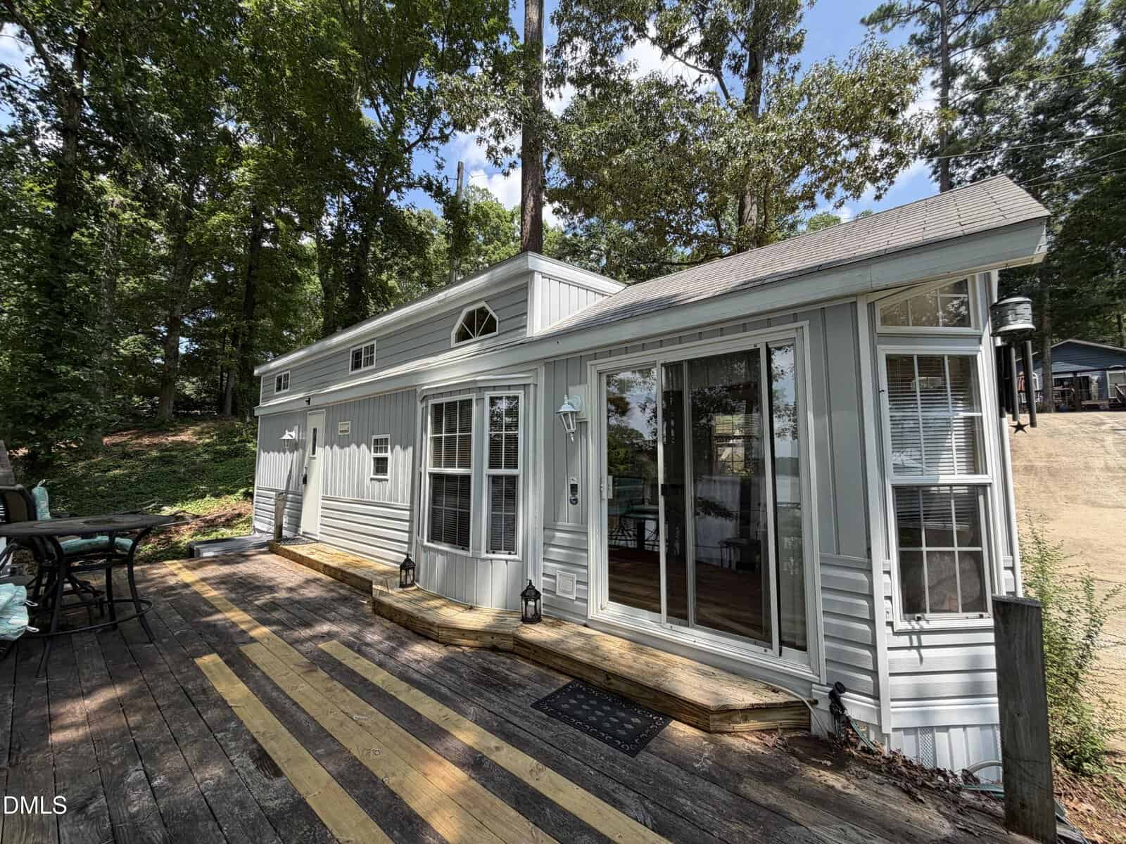 Small, light-gray house with large windows and a sloped roof, surrounded by trees. Wooden deck with a seating area in front. Sunny day with blue sky and scattered clouds.