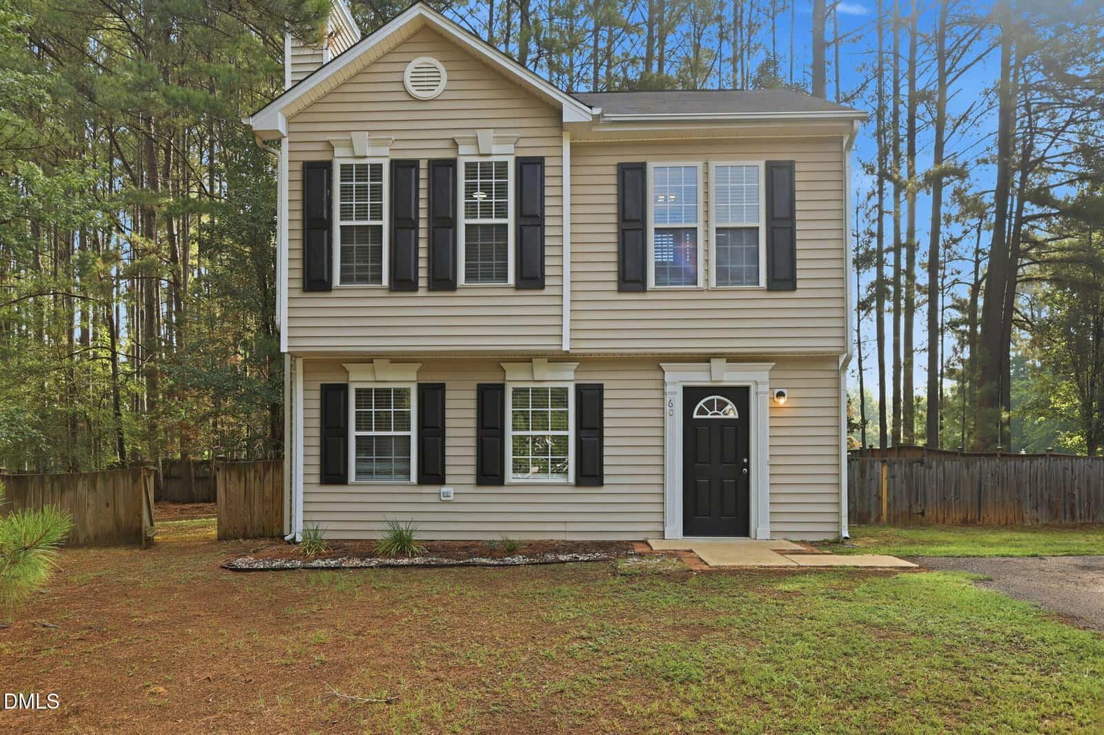 Two-story beige house with black shutters, white trim, and a black front door located at 60 Holding-Young Road in Youngsville. Surrounded by tall pine trees and a wooden fence, this home features a simple front yard and a small garden bed near the entrance.