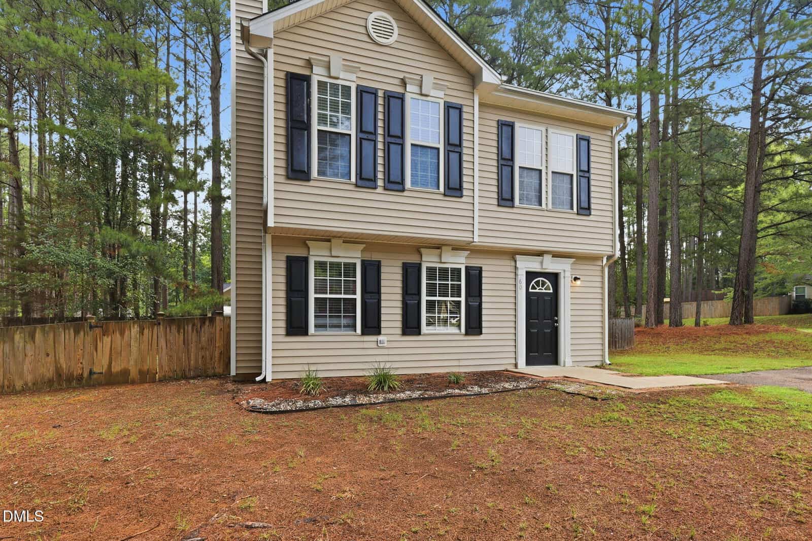 Two-story beige house with black shutters and a black front door, surrounded by tall pine trees, a wooden fence, and a brown mulched yard with a small concrete path leading to the entrance.