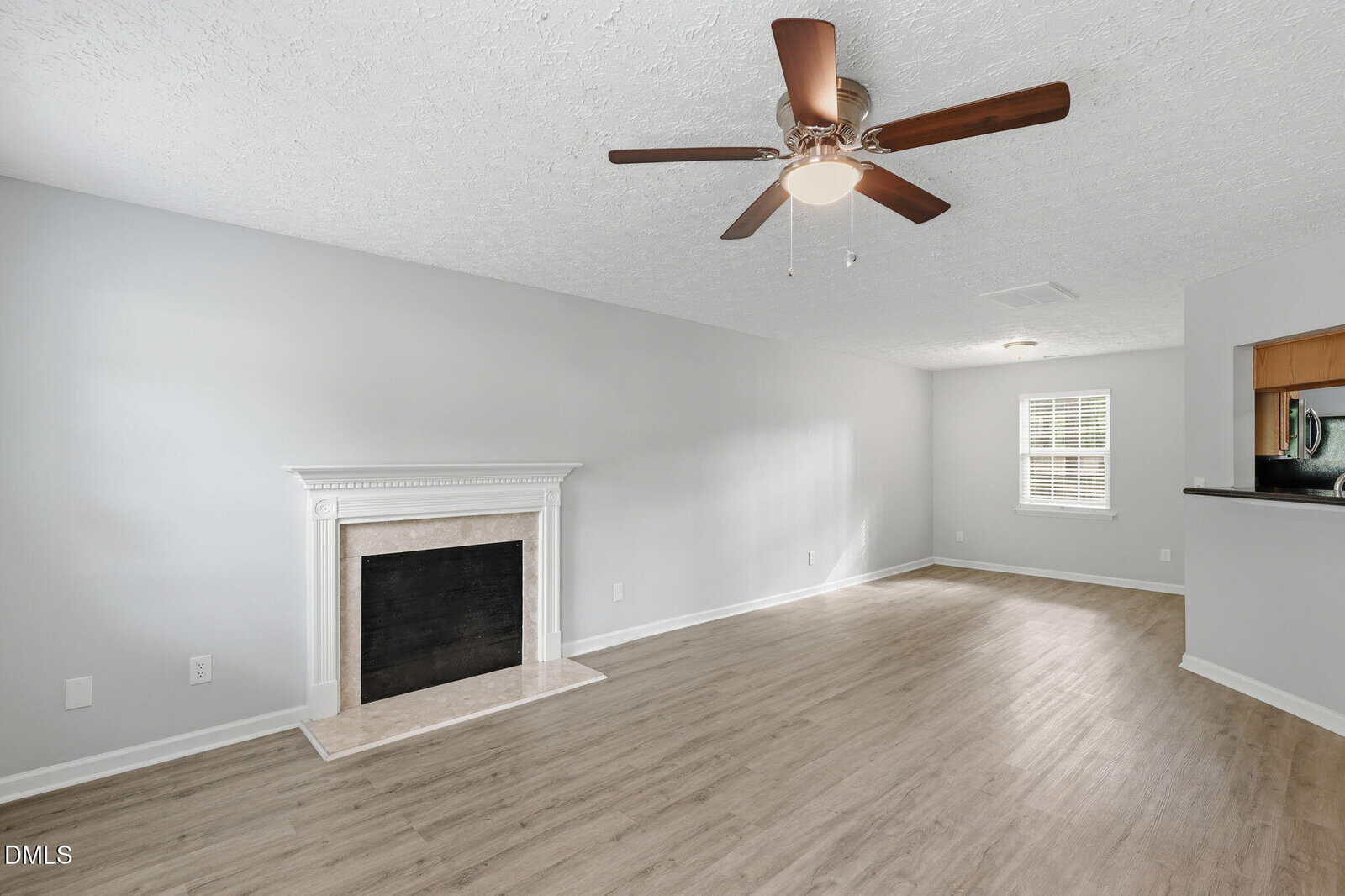 A bright, empty living room features light wood floors, a white fireplace, gray walls, a ceiling fan, and a large window with blinds. An open kitchen area is visible on the right.