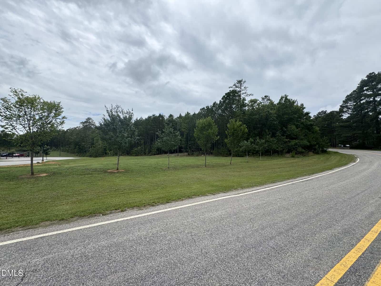 A curved paved road bordered by green grass and young trees leads past a wooded area under a cloudy sky.