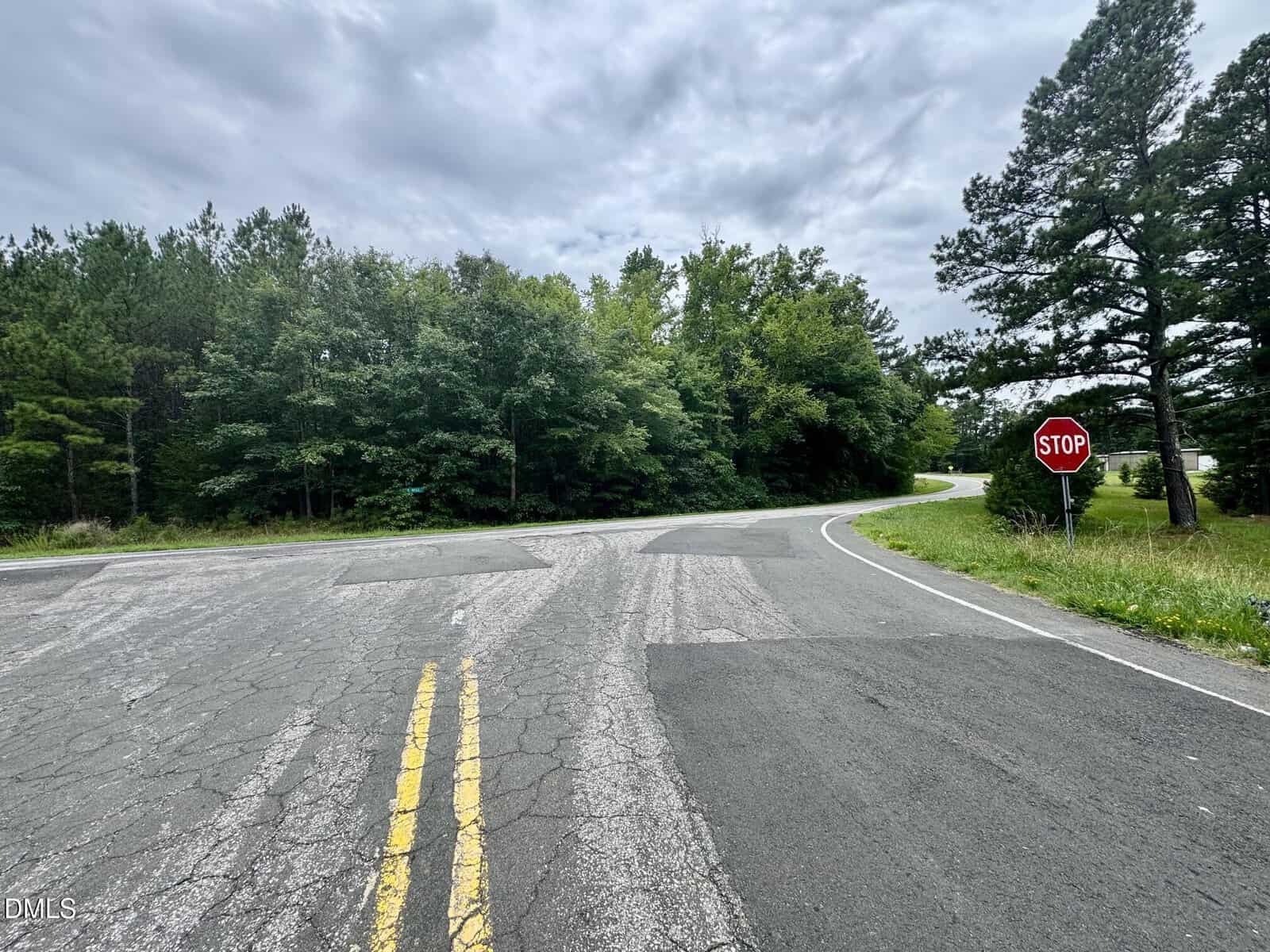 A cracked rural road ends at a T-intersection bordered by green trees. A red stop sign stands on the right. The sky is cloudy, and the road curves to the right beyond the intersection.