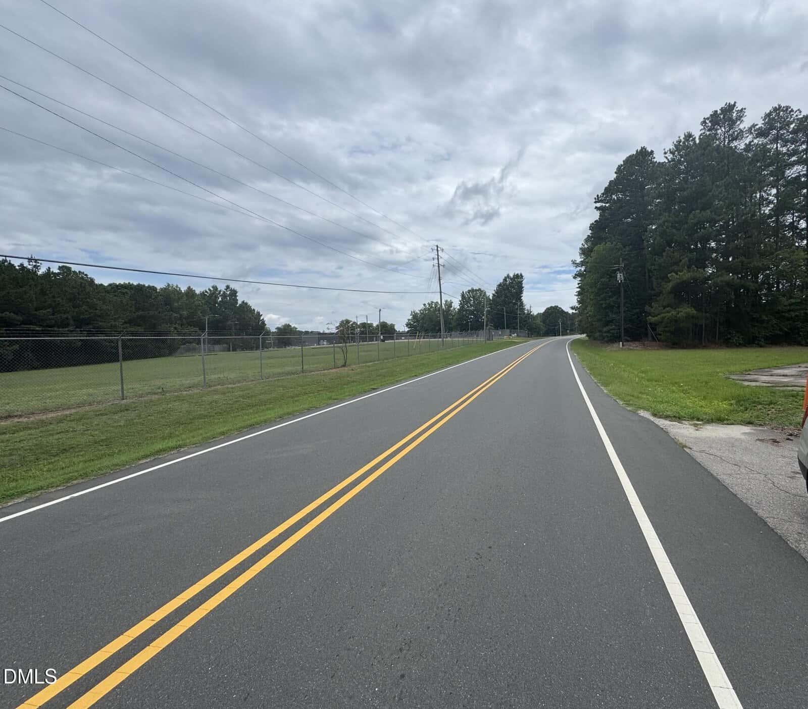 A two-lane road with double yellow lines curves gently right, bordered by grassy areas, trees, and a chain-link fence under a cloudy sky. No vehicles or people are visible.