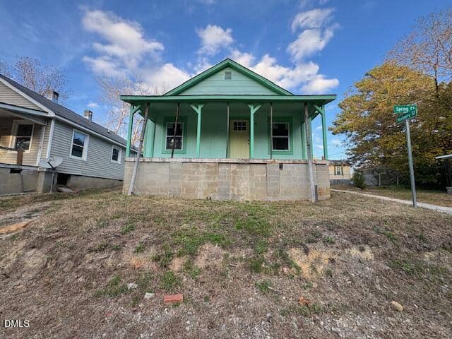 A small green house with a front porch sits on a raised concrete foundation at the corner of Spring Street in Henderson, with patches of grass and bare ground in the yard under a partly cloudy sky.