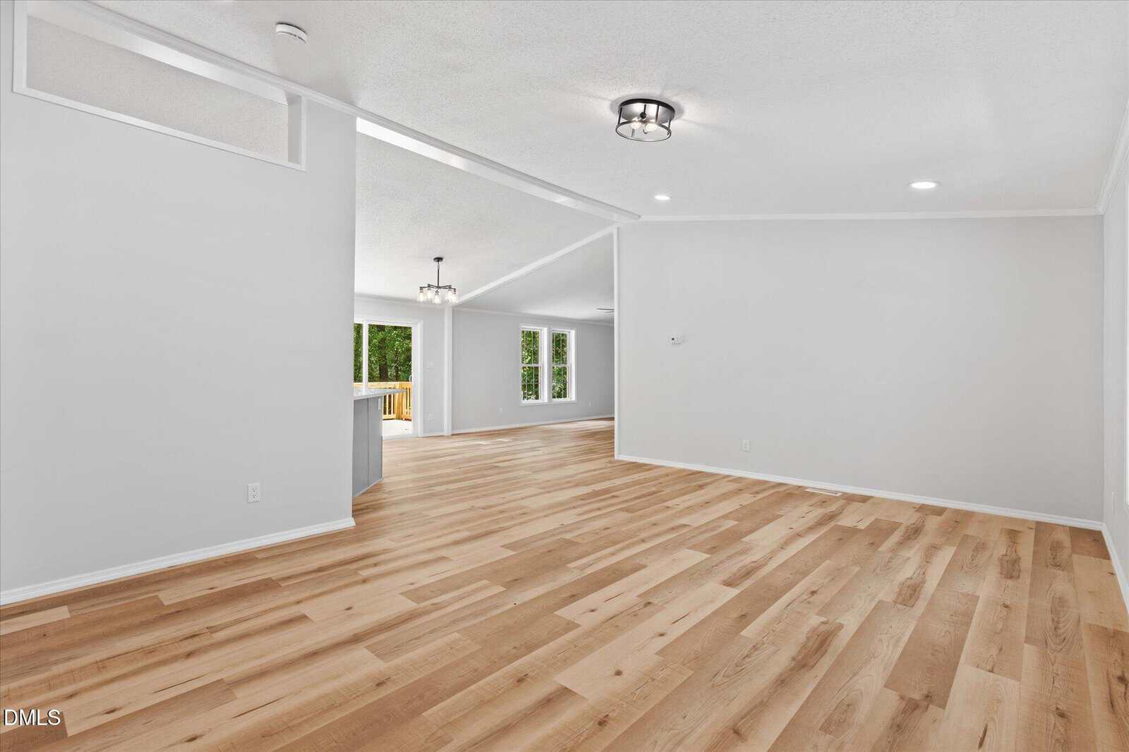 Spacious, empty room with light wood floors, white walls, and ceiling lights. Large windows and open doorway lead to a bright kitchen and dining area in the background.