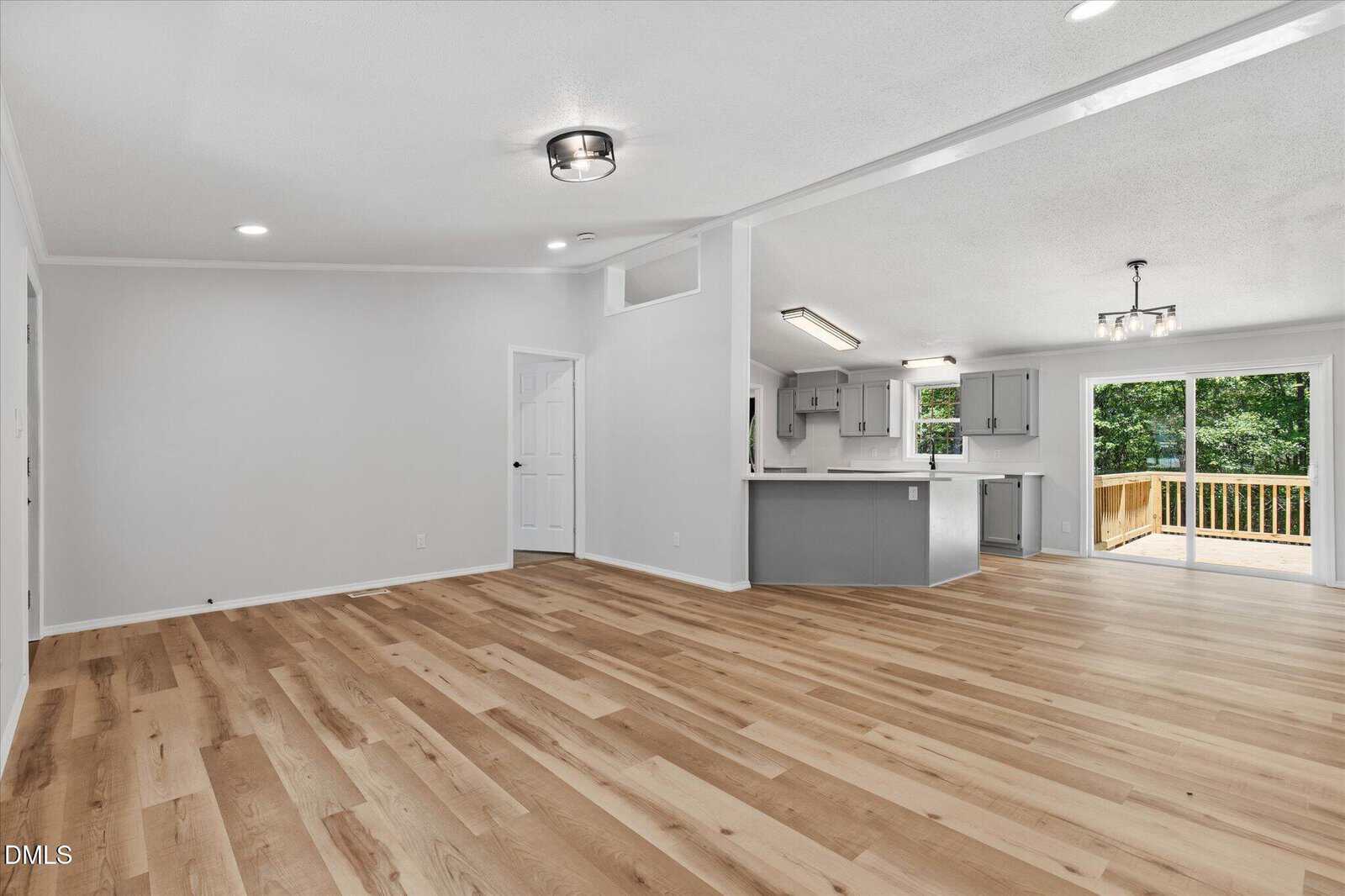Open-concept living area with light wood flooring, white walls, a modern kitchen with gray cabinets, and sliding glass doors leading to a wooden deck outside. Natural light fills the space.