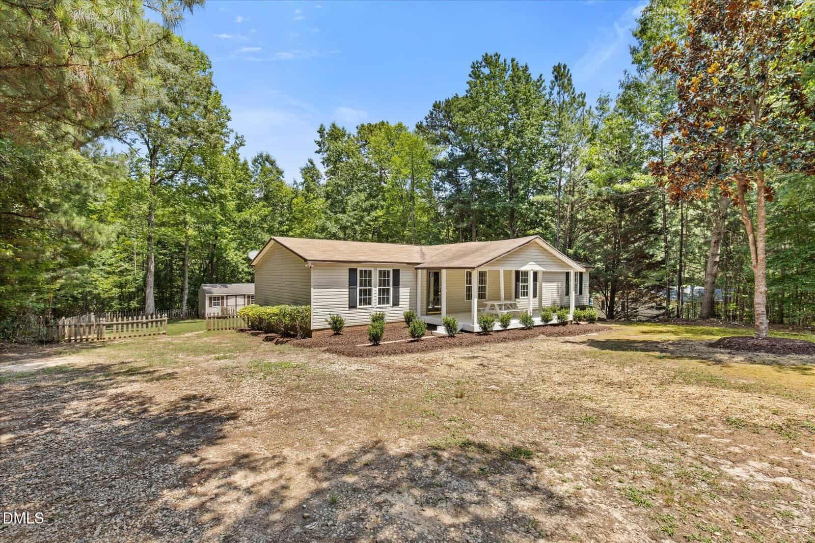Single-story beige house with a front porch, white railings, and flower beds, nestled among trees on a spacious yard at 175 Windsong Lane in Youngsville, all beneath a clear blue sky.