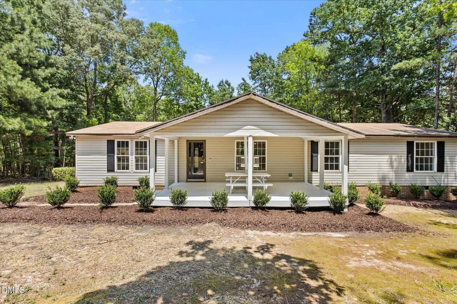 Single-story beige house with a covered front porch, white railings, and shutters, surrounded by green trees and neatly trimmed bushes, on a dirt and grass yard under a clear blue sky.