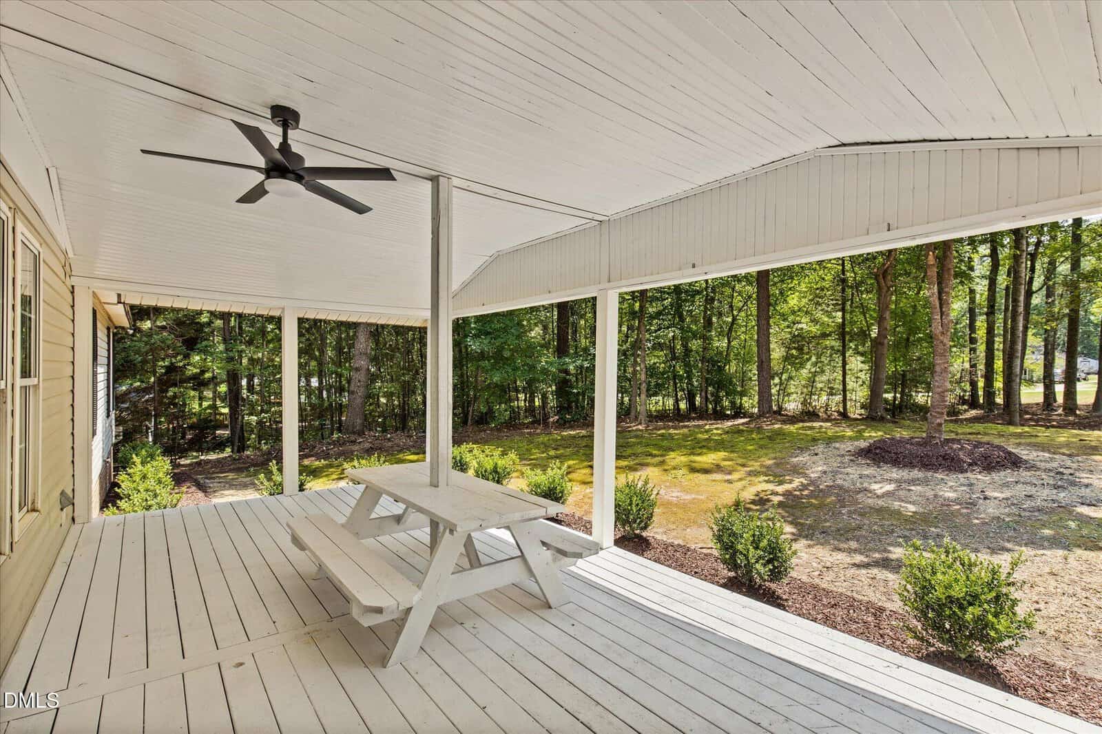 A white wooden porch with a picnic table, ceiling fan, and view of a grassy yard bordered by trees and shrubs on a sunny day.