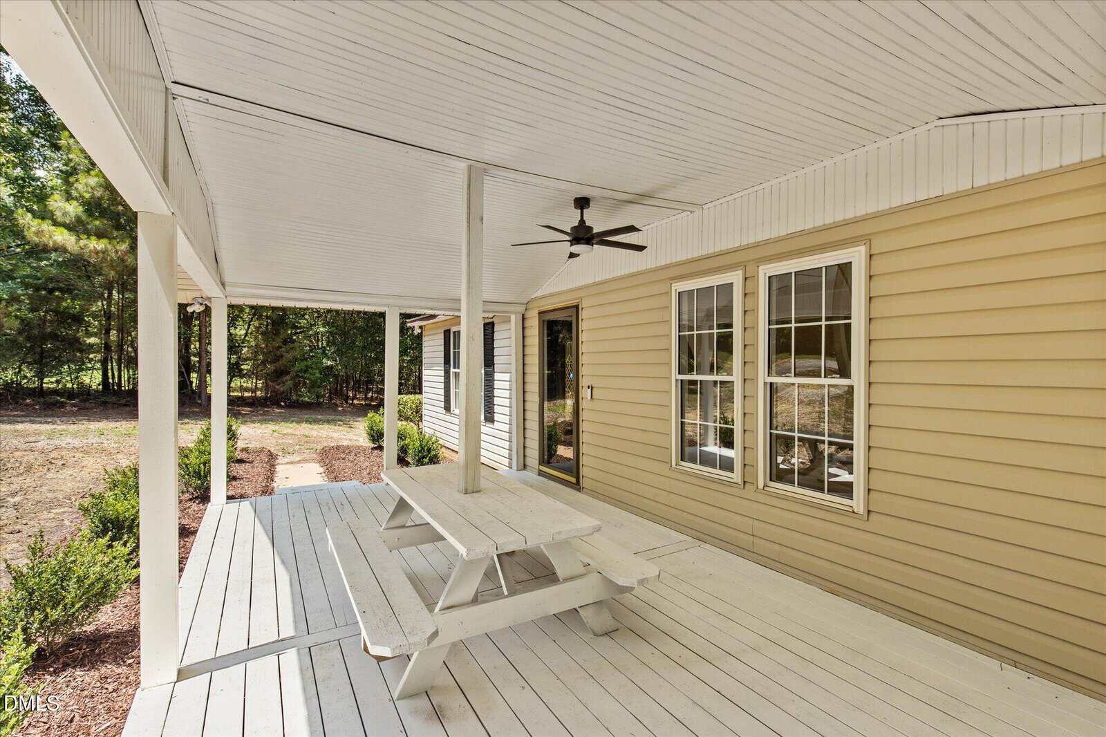 Covered porch with white flooring and ceiling, yellow siding, double windows, black ceiling fan, and a white picnic table. Trees and bushes are visible in the background.