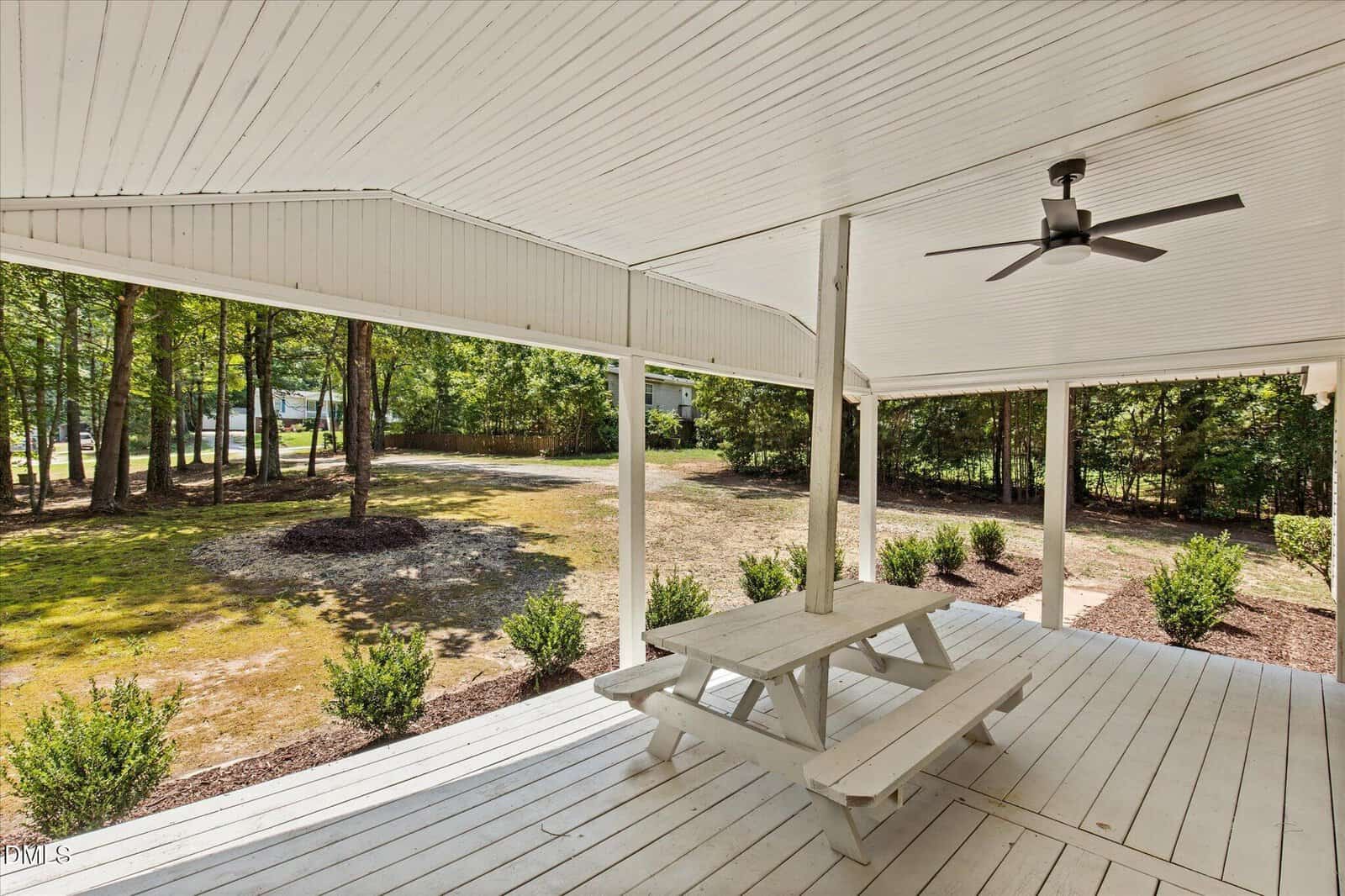 A covered white wooden porch with a ceiling fan and a white picnic table overlooks a green yard with trees and bushes. The porch has open sides, letting in natural light.