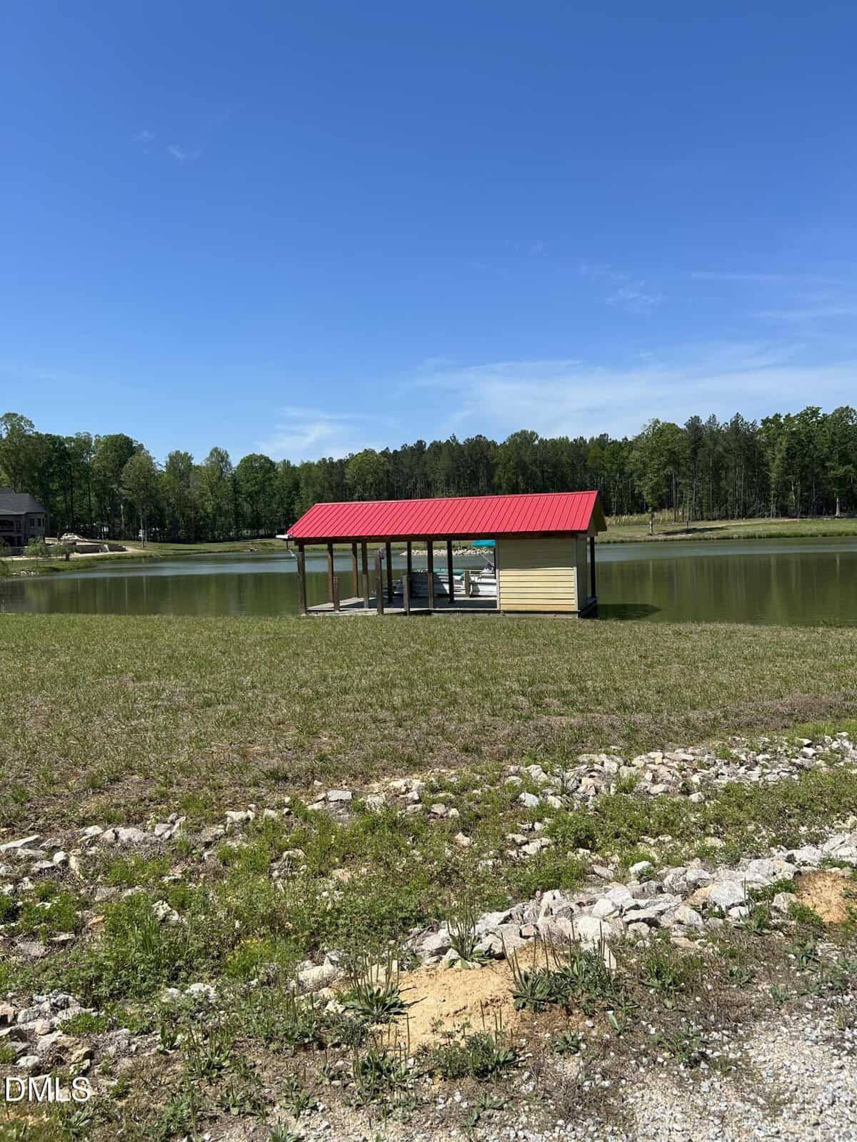 A small wooden dock with a red roof sits on a calm lake, surrounded by grassy land and trees under a clear blue sky.