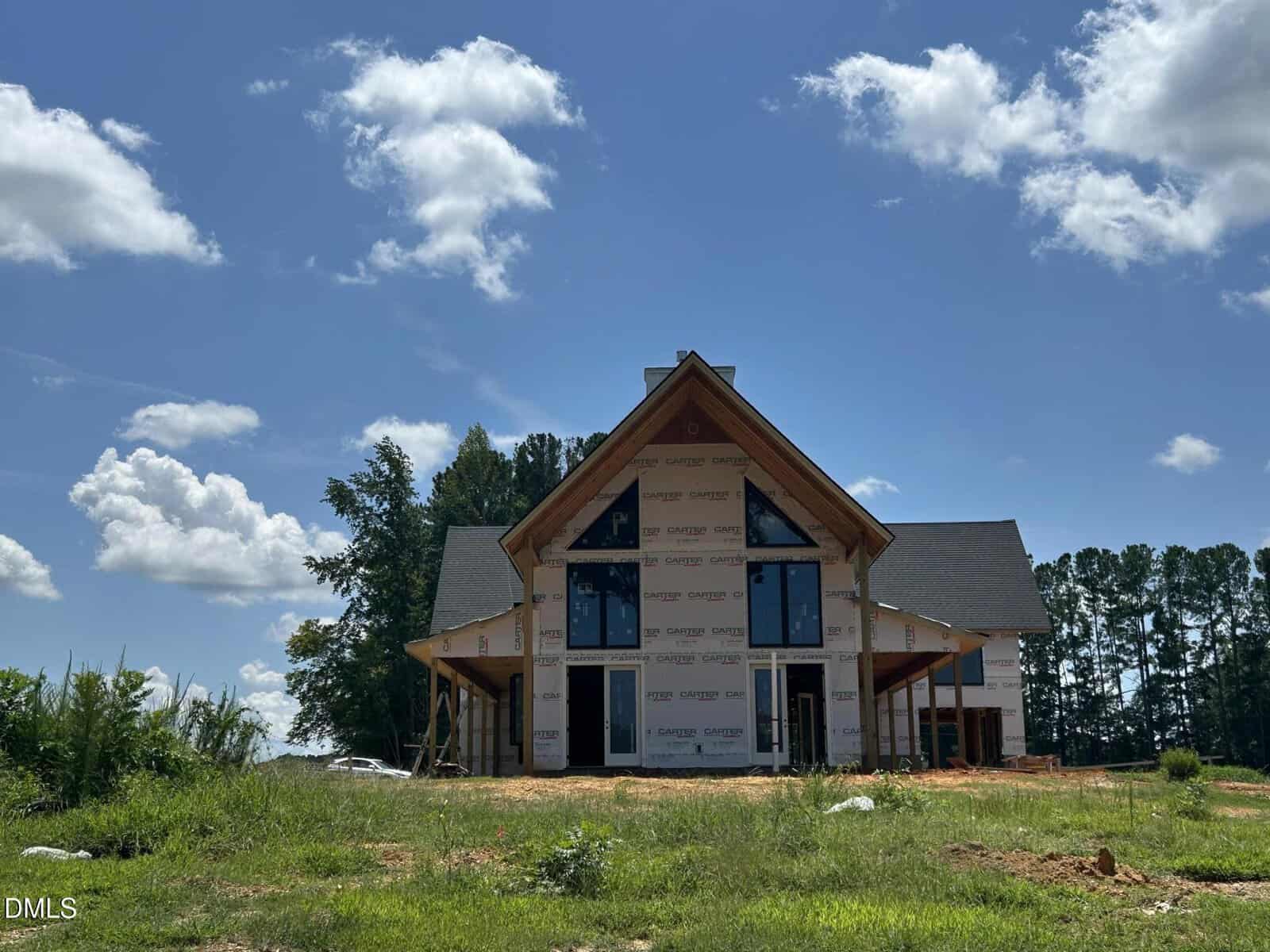 A partially constructed house with visible framing and house wrap stands on a dirt lot, surrounded by grass and trees, under a blue sky with scattered clouds.