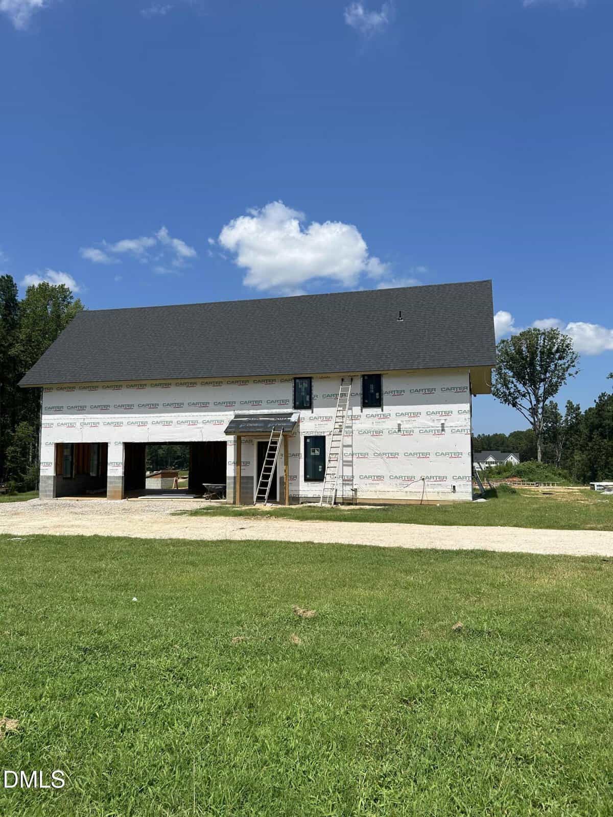 A two-story house under construction with black roof shingles and white weatherproofing wrap, ladders leaning against the front, and a grassy yard under a clear blue sky.
